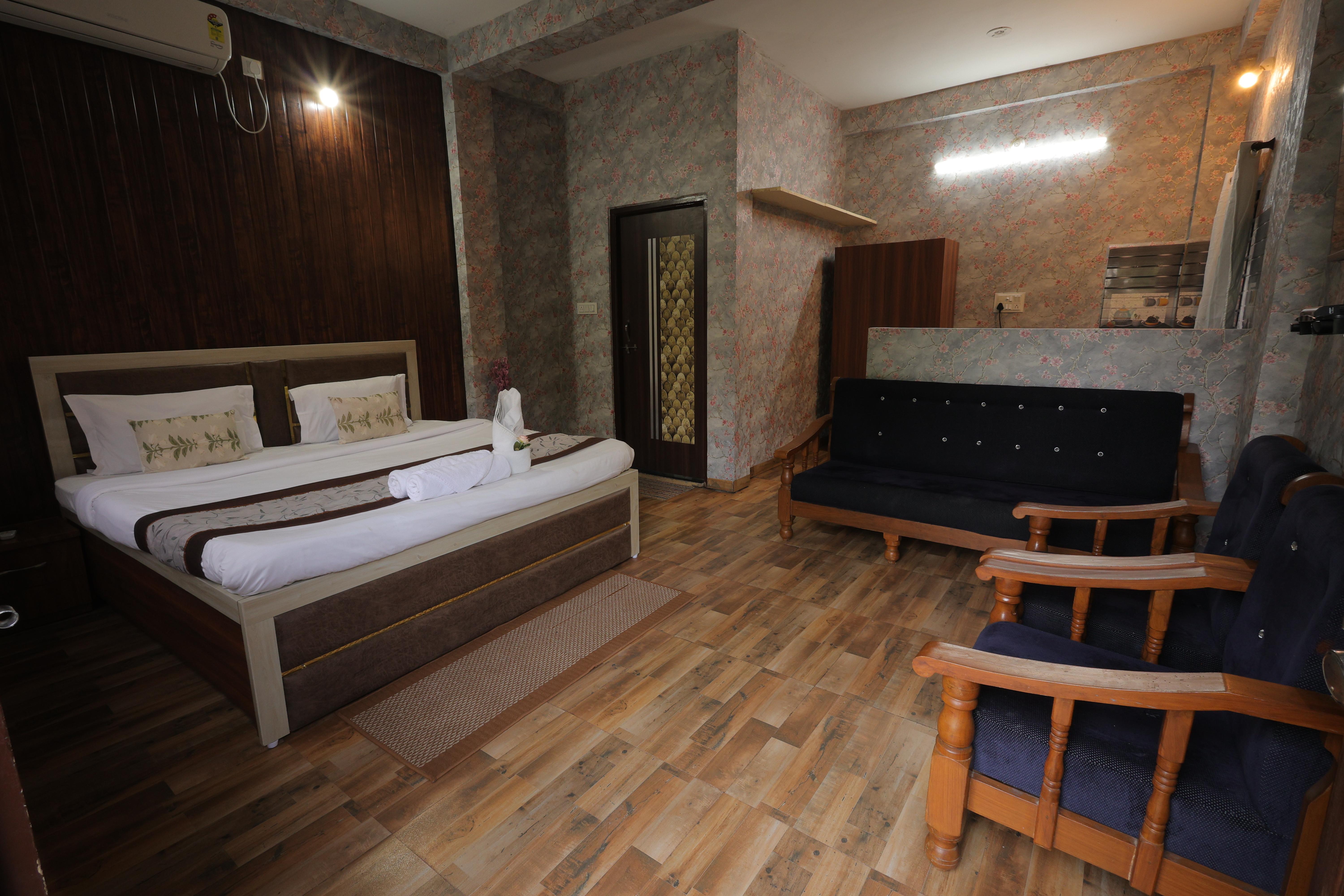 A cozy and elegant villa room at Nature Nest Hotel &amp; Resorts, designed for comfort and relaxation. The room features a luxurious king-sized bed with neatly arranged linens and decorative pillows, creating a warm and inviting ambiance. The wooden flooring and earthy-toned decor complement the rustic yet modern aesthetic.  A comfortable seating area with a black cushioned sofa and wooden armchairs adds to the charm, making it perfect for lounging. The soft lighting, floral-patterned wallpaper, and well-organized space enhance the peaceful retreat experience, offering a blend of nature-inspired elegance and modern comfort.
