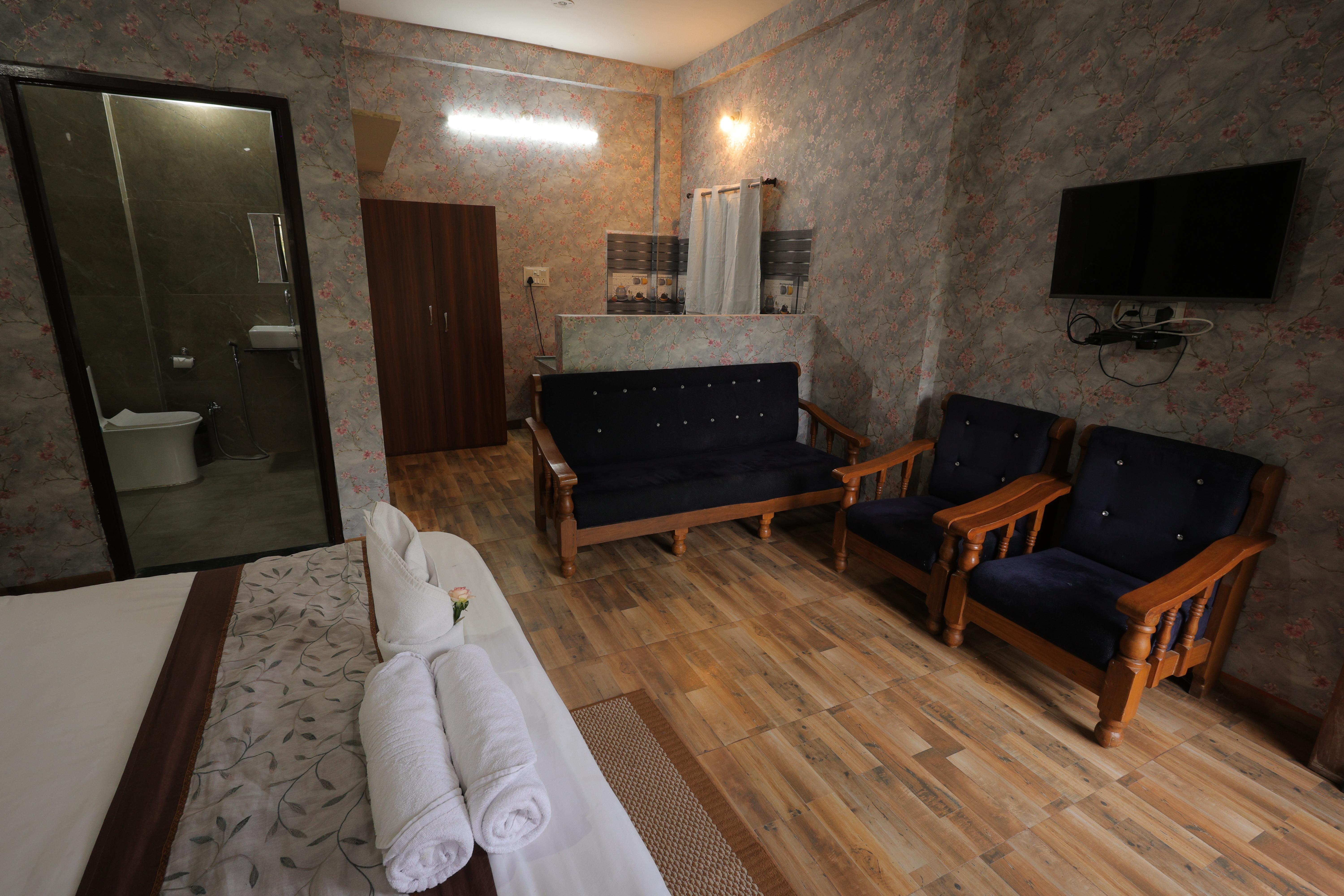 A spacious and elegantly designed villa at Nature Nest Hotel &amp; Resorts, perfect for a cozy and luxurious stay. The room features a comfortable king-size bed with neatly arranged fresh white towels, ensuring a relaxing experience.  The wooden sofa set with plush navy blue cushions adds a touch of sophistication and provides a perfect space to unwind. The wall-mounted TV ensures entertainment at your fingertips. The attached bathroom with modern fittings, a wardrobe for storage, and a kitchenette with essential amenities make this villa an ideal retreat for a comfortable and peaceful getaway.