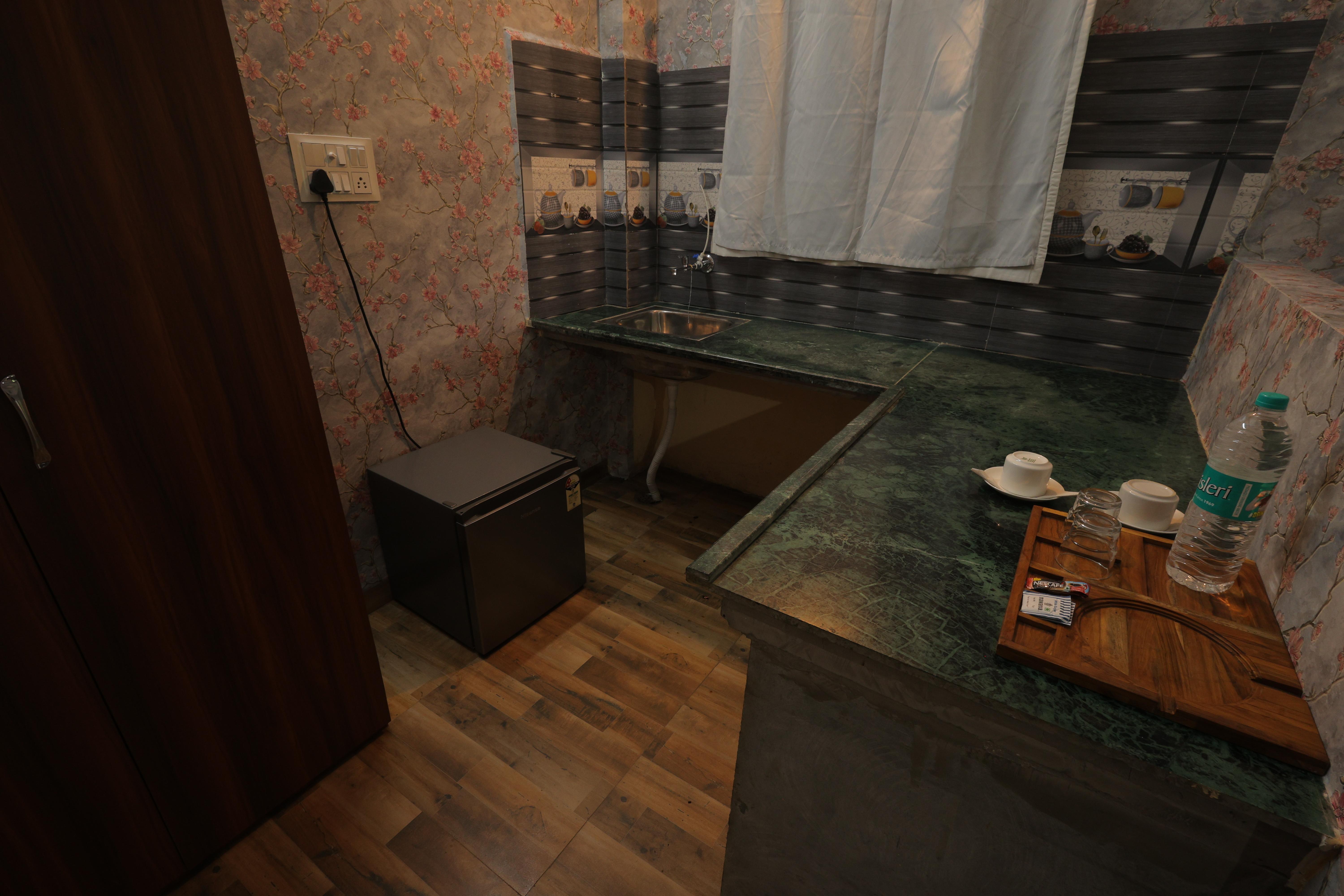 A compact yet well-equipped kitchenette inside a villa at Nature Nest Hotel &amp; Resorts. The space features green marble countertops, a stainless steel sink, and a mini refrigerator for added convenience. The wooden flooring and floral wallpaper enhance the cozy ambiance, making it a great spot for guests to prepare light meals or enjoy a warm cup of tea.  A wooden tray with tea cups, glasses, and bottled water is thoughtfully arranged for guests. The wall-mounted tiles with a tea and coffee theme add a charming touch to the space. Whether you're looking for a quick refreshment or a peaceful moment with your favourite brew, this kitchenette offers a perfect retreat. 