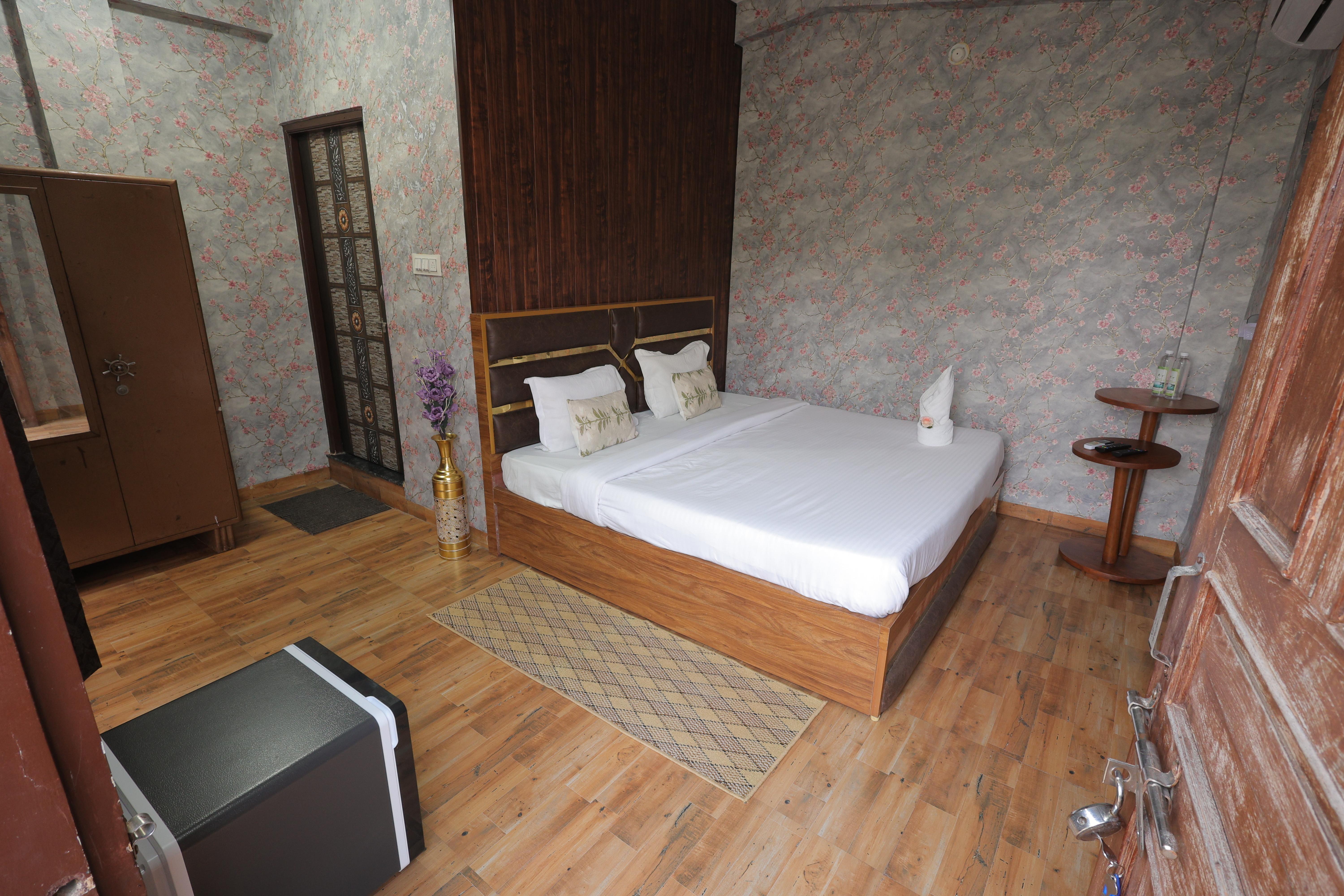 A cozy and elegantly designed villa room at Nature Nest Hotel &amp; Resorts, offering a warm and inviting ambiance. The room features a comfortable wooden bed with crisp white linens and floral-themed cushions, enhancing its rustic charm. The wooden flooring and subtle floral wallpaper create a perfect blend of nature and comfort.  A small wooden side table holds bottled water and essentials, ensuring a convenient stay. The classic wooden wardrobe and decorative elements, such as the stylish vase with flowers, add a touch of sophistication. The soft lighting and earthy tones make this villa room an ideal retreat for those looking to relax and reconnect with nature. 