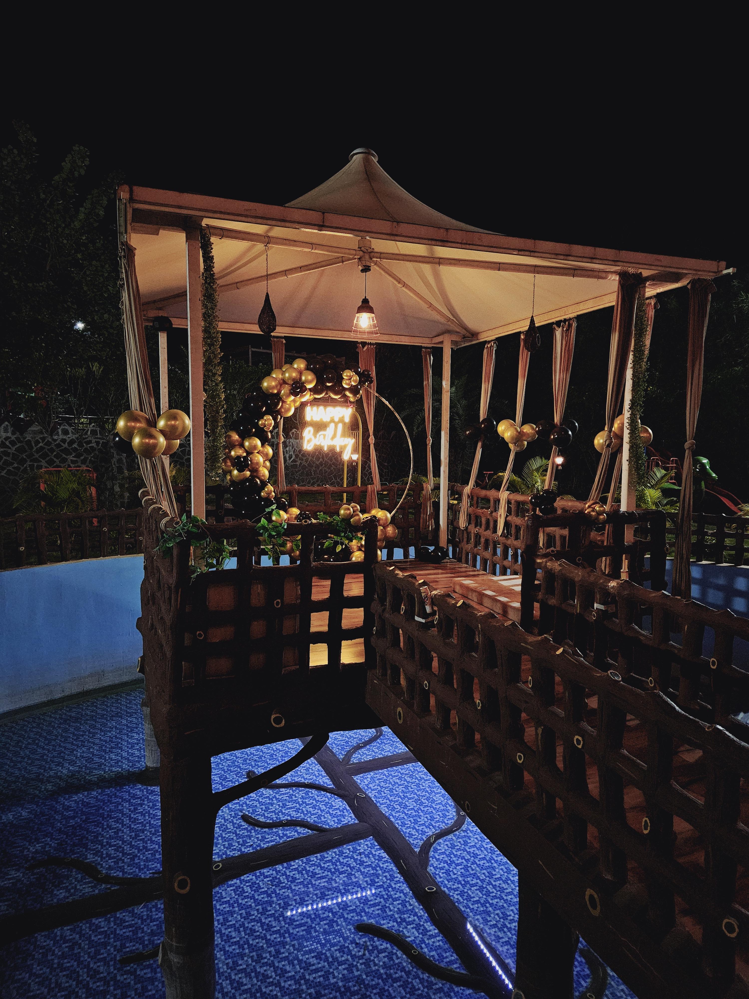 A dreamy birthday celebration under the stars! This stunning gazebo setup, glowing with warm lights and golden-black balloons, creates the perfect ambiance for a magical evening. Surrounded by nature and floating above a beautifully lit pool, this is the ultimate way to celebrate in style.