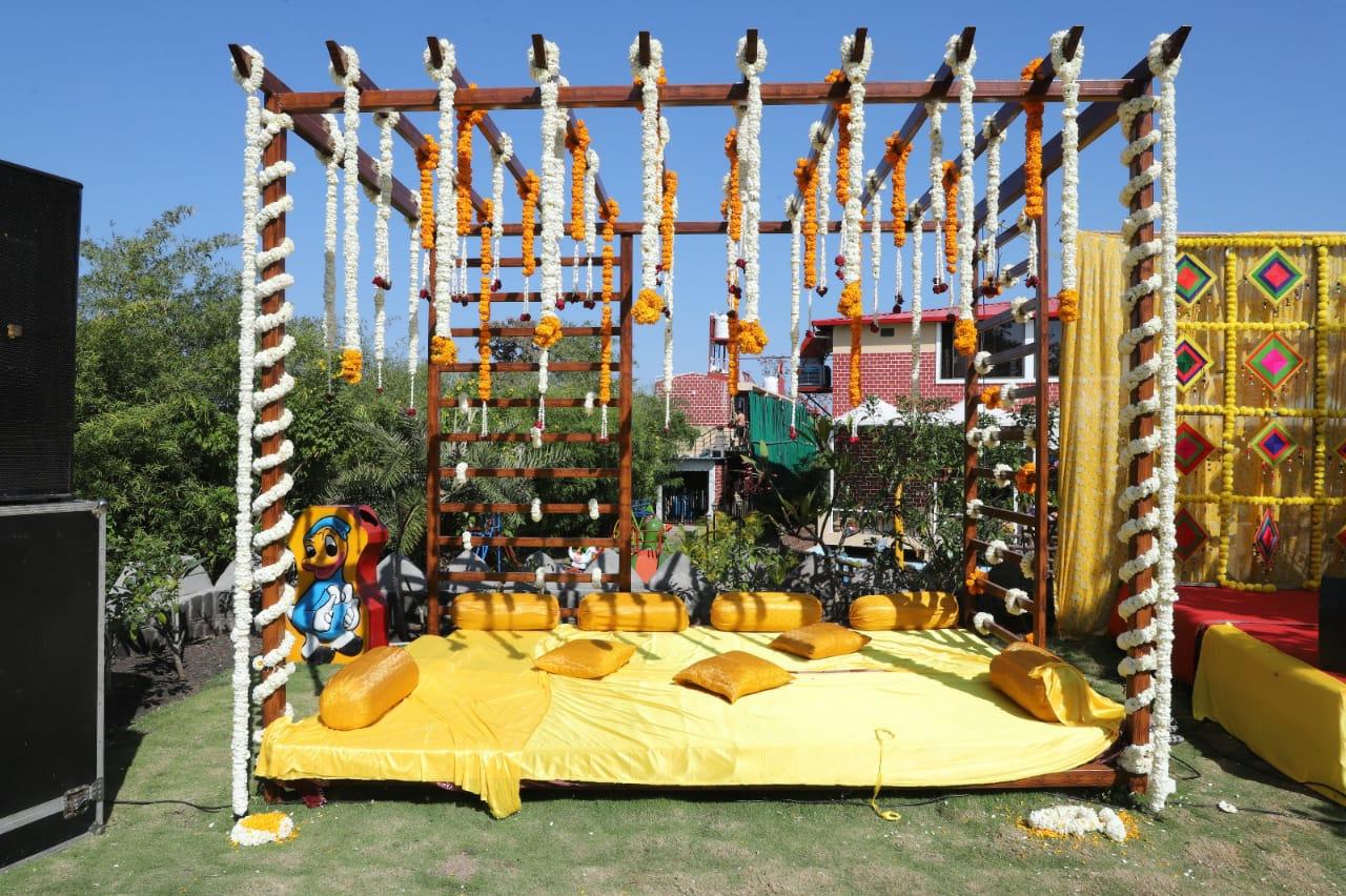 A beautifully crafted outdoor seating arrangement for a Haldi ceremony, featuring a wooden pergola adorned with marigold and jasmine garlands. The vibrant yellow cushions and draped fabric create a cozy and festive atmosphere, perfect for traditional pre-wedding celebrations. The open-air setting, surrounded by lush greenery, enhances the natural charm, making it an ideal spot for a joyful and intimate gathering