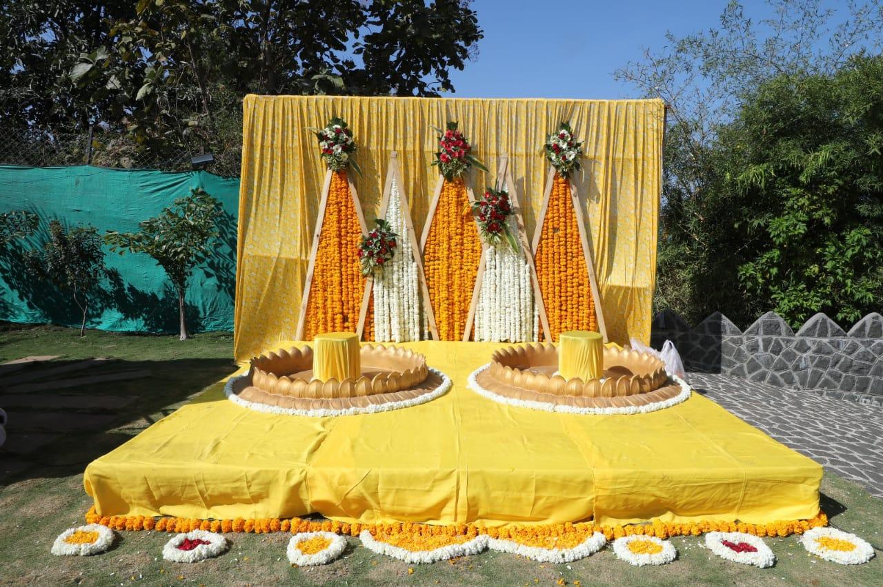 A stunning Haldi ceremony setup featuring a vibrant yellow and orange floral theme, set against a lush outdoor backdrop. The stage is adorned with traditional marigold garlands, elegant floral arrangements, and decorative elements that add a touch of grace and festivity. The symmetrical design with golden seating arrangements enhances the charm, making it a perfect spot for a joyful pre-wedding celebration amidst nature.