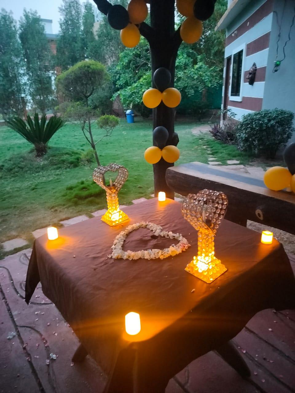 A romantic candlelit setup with warm golden hues, heart-shaped decor, and a charming black-and-yellow theme. The cozy outdoor ambiance, surrounded by lush greenery, makes it a perfect setting for a special evening.